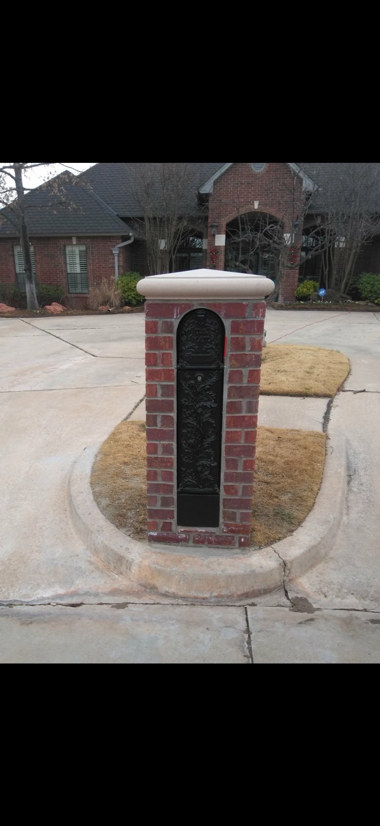 Red brick mailbox with white cap on residential driveway, brick house visible in background